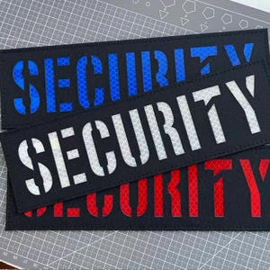 Op de afbeelding: Drie rechthoekige zwarte patches met het woord SECURITY in dikke hoofdletters. De bovenste patch heeft blauwe reflecterende tekst, de middelste patch heeft witte reflecterende tekst en de onderste patch heeft rode reflecterende tekst.