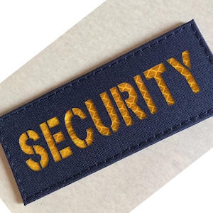 May include: A navy blue rectangular patch with the word "SECURITY" in yellow, reflective lettering. The patch has a stitched border and is likely for uniform identification.