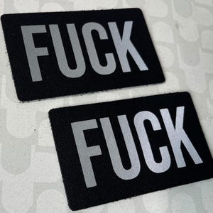 Op de afbeelding: Twee zwarte rechthoekige patches met het woord "FUCK" in grote, zilveren blokletters. De patches zijn gemaakt van een textuurrijk materiaal en worden getoond tegen een witte achtergrond.