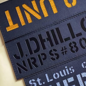 May include: Close-up of several navy blue rectangular patches with cut-out lettering. The top patch reads "UNIT" in yellow, while the others feature black and white text, including "J.DHILL", "NRPS #80", and "St. Louis".