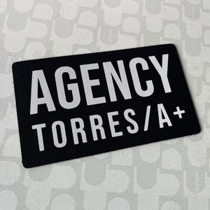 May include: A black rectangular patch with the white text "AGENCY TORRES/A+". The patch is set against a light grey background with a repeating pattern.