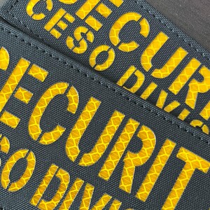 Peut inclure: Deux &eacute;cussons rectangulaires en tissu noir portant le mot SECURITY en lettres capitales d&eacute;coup&eacute;es. Le lettrage r&eacute;v&egrave;le un mat&eacute;riau r&eacute;fl&eacute;chissant jaune vif &agrave; motif en nid d'abeille sous la surface sombre et textur&eacute;e des &eacute;cussons.