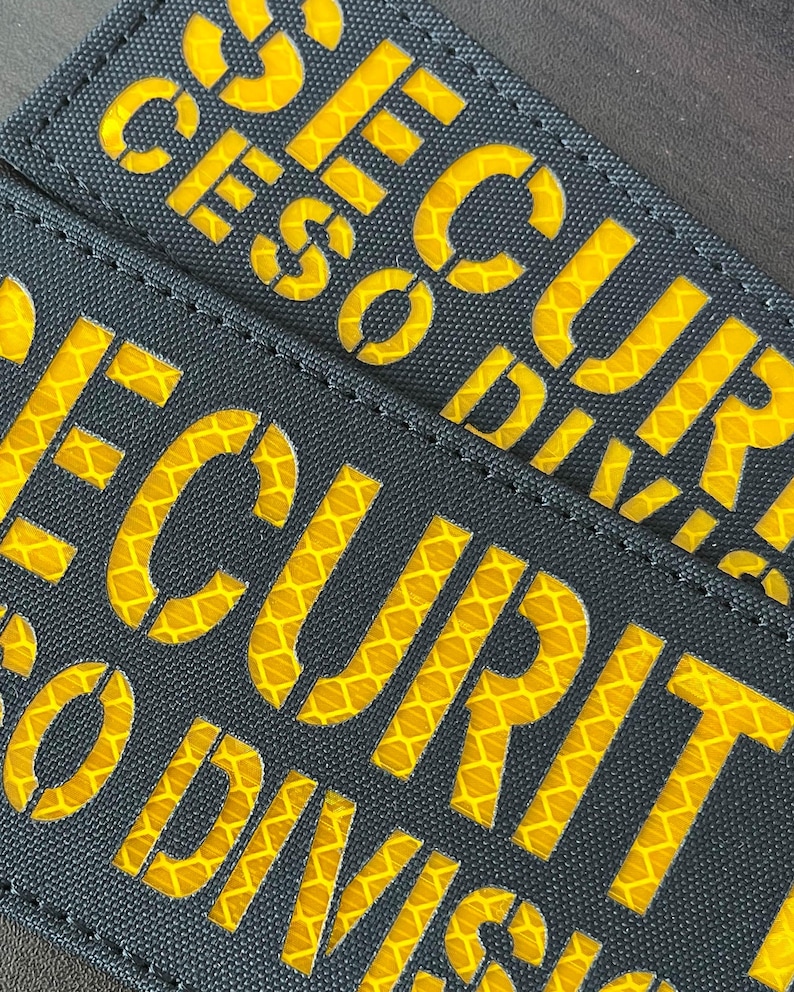 Peut inclure: Deux &eacute;cussons rectangulaires en tissu noir portant le mot SECURITY en lettres capitales d&eacute;coup&eacute;es. Le lettrage r&eacute;v&egrave;le un mat&eacute;riau r&eacute;fl&eacute;chissant jaune vif &agrave; motif en nid d'abeille sous la surface sombre et textur&eacute;e des &eacute;cussons.
