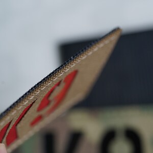 May include: Close-up of a tan and black patch with red stitching. The patch has a black border and a red design.