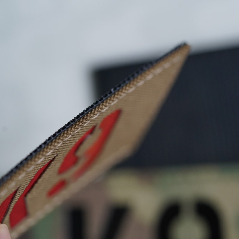 May include: Close-up of a tan and black patch with red stitching. The patch has a black border and a red design.