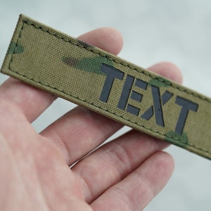 以下が含まれることがあります： 黒いブロック体の「TEXT」という文字が入った長方形のパッチ。パッチは、緑、茶色、タンの色合いのカモフラージュ柄です。縁は濃い緑色の糸でステッチされています。