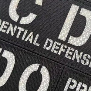 May include: A black rectangular patch featuring reflective white lettering with a honeycomb pattern. The visible text includes the letters C and D, the word ENTIAL, the word DEFENSE, the letters D and O, and the letters PR.