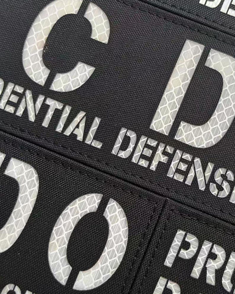 May include: A black rectangular patch featuring reflective white lettering with a honeycomb pattern. The visible text includes the letters C and D, the word ENTIAL, the word DEFENSE, the letters D and O, and the letters PR.
