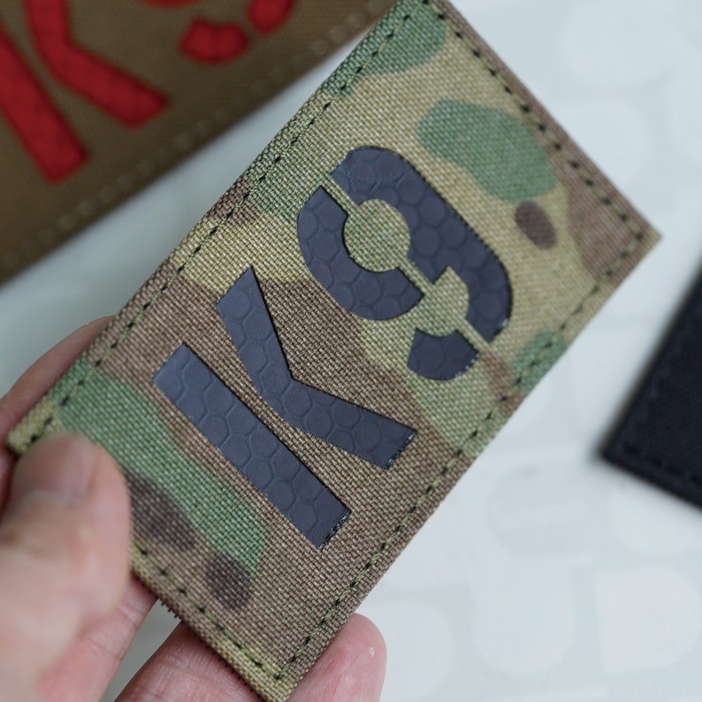 May include: A black K9 patch with a camouflage background. The patch is sewn with black thread around the edges.