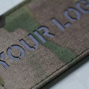 K&ouml;nnte beinhalten: Ein rechteckiger Stoffaufn&auml;her mit einem Tarnmuster in Gr&uuml;n-, Braun- und Beiget&ouml;nen. Der Aufn&auml;her weist lasergeschnittene Buchstaben auf, die YOUR LOGO bilden und eine dunkle, solide Hintergrundschicht unter der strukturierten Textiloberfl&auml;che zeigen.