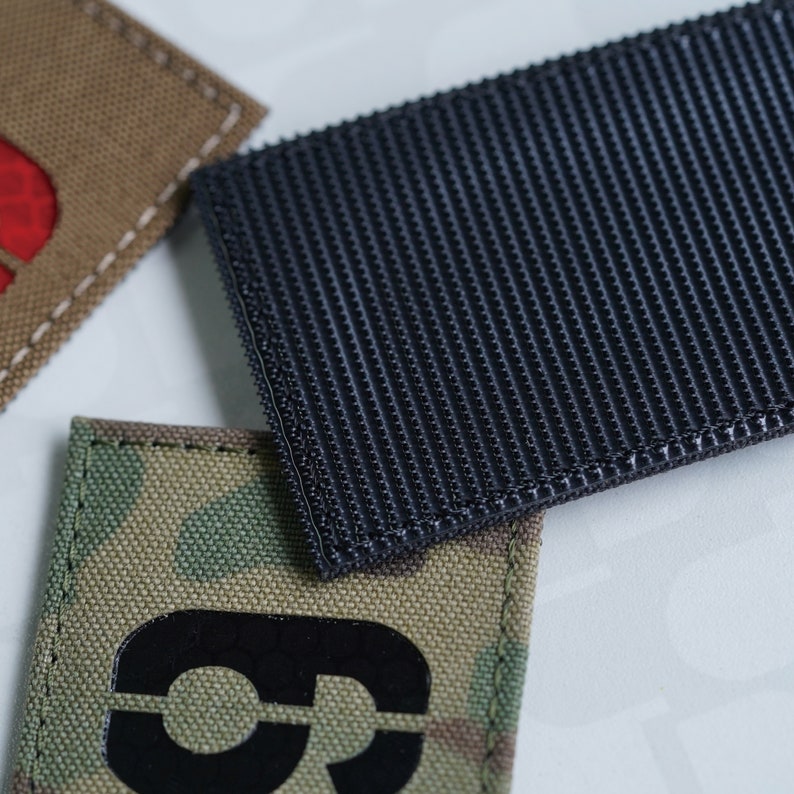 May include: A camouflage fabric patch with a black number "3" in the center. The patch is attached to a black hook and loop fastener.