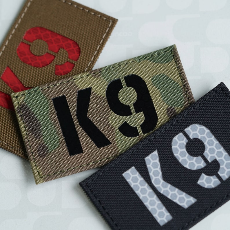 May include: Three K9 patches with different backgrounds and reflective lettering. The first patch is brown with red reflective lettering. The second patch is camouflage with black reflective lettering. The third patch is black with white reflective lettering.
