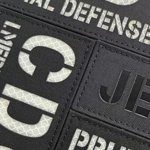 May include: A black rectangular patch featuring reflective white lettering that reads "INITIAL DEFENSE" at the top, with partial letters "CD" visible below and "PRUD" visible at the bottom, alongside a separate black patch with the black text "JE".