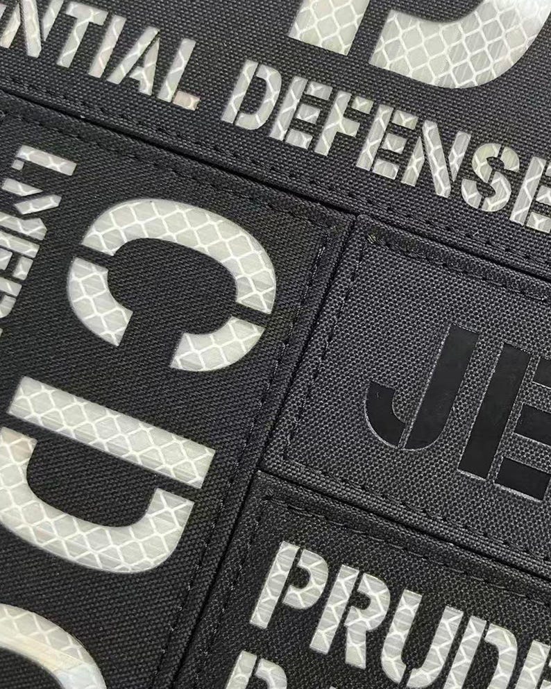 May include: A black rectangular patch featuring reflective white lettering that reads "INITIAL DEFENSE" at the top, with partial letters "CD" visible below and "PRUD" visible at the bottom, alongside a separate black patch with the black text "JE".