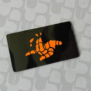 May include: Rectangular patch with a camouflage background and an orange skeletal hand design. The patch has a black border and is likely made of durable fabric. The design is centered on the patch.
