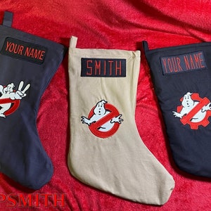 Op de afbeelding: Drie zwarte en beige kerstsokken met een Ghostbusters-logo en gepersonaliseerde naamlabels. De sokken hebben een rood 'geen geest'-symbool met een witte geest erin. De naamlabels luiden 'Uw naam', 'Smith' en 'Uw naam'.