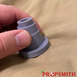 May include: A gray plastic prop replica of a Ghostbusters proton pack nozzle. The nozzle is made up of several stacked rings and has a smooth, rounded surface.