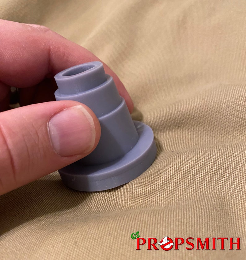 May include: A gray plastic prop replica of a Ghostbusters proton pack nozzle. The nozzle is made up of several stacked rings and has a smooth, rounded surface.