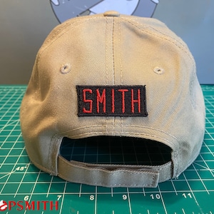 Op de afbeelding: Een beige baseballpet met een zwart en rood geborduurd patch met de tekst "SMITH" op de achterkant.