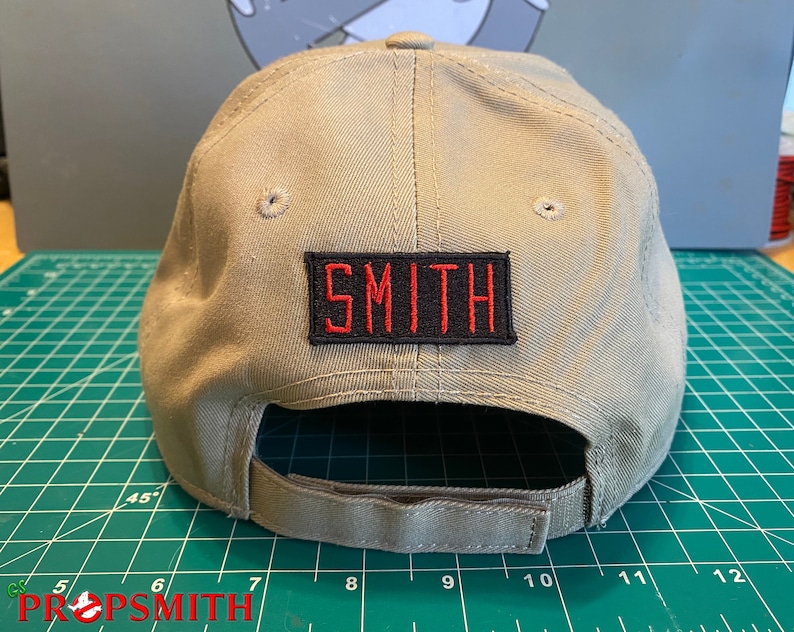 Op de afbeelding: Een beige baseballpet met een zwart en rood geborduurd patch met de tekst "SMITH" op de achterkant.