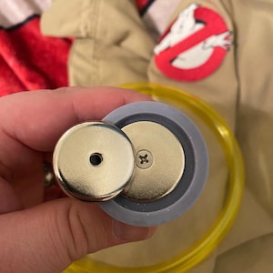 May include: Close-up of a silver-colored, round, magnetic button with a small hole in the center. The button is attached to a gray, round, plastic base with a screw in the center. The button is being held by a hand.