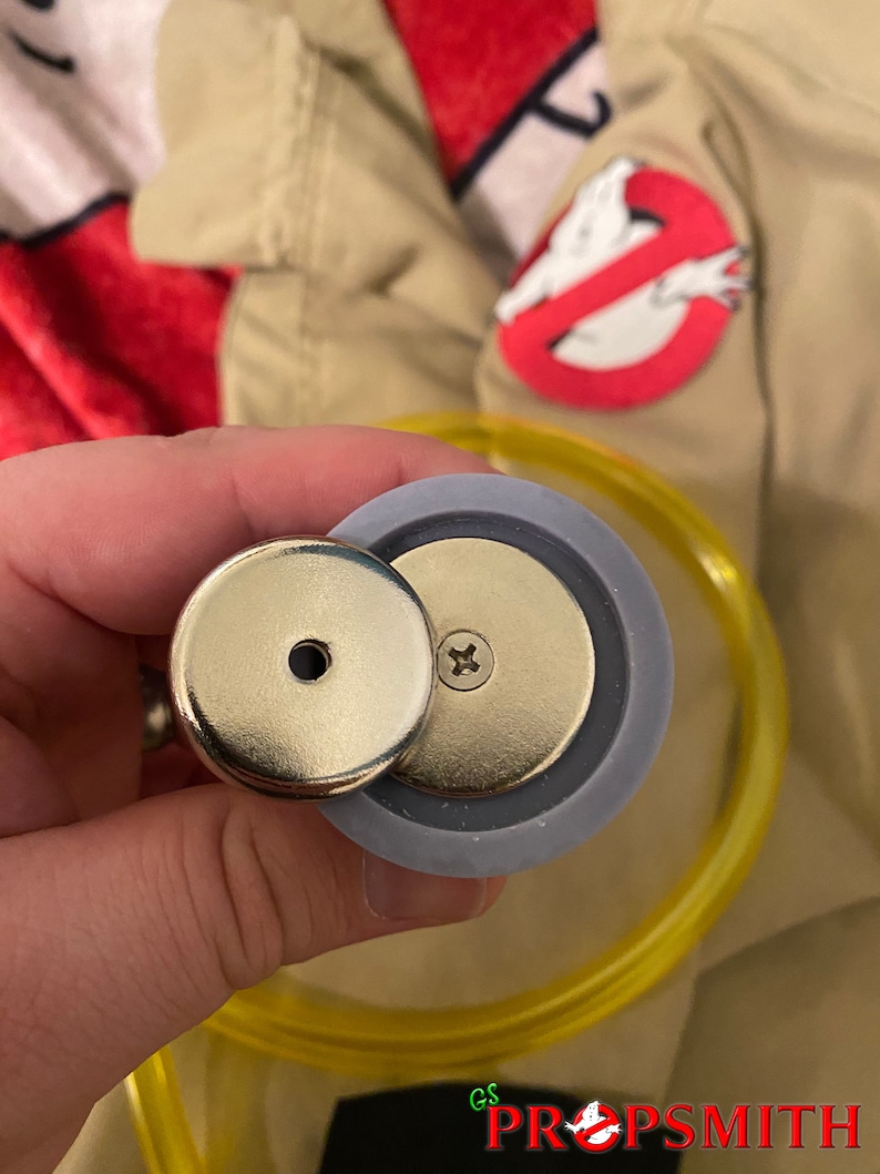 May include: Close-up of a silver-colored, round, magnetic button with a small hole in the center. The button is attached to a gray, round, plastic base with a screw in the center. The button is being held by a hand.