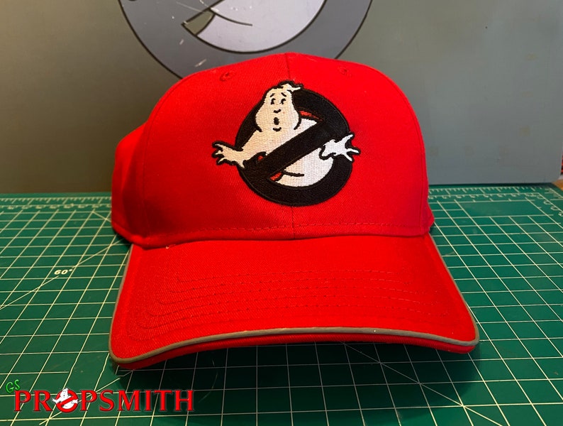 Op de afbeelding: Rode baseballpet met een geborduurd Ghostbusters-logo in zwart en wit met het 'geen geest'-symbool en een cartoon-geest.