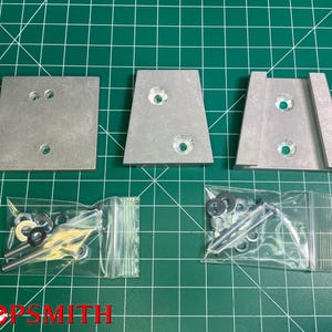 Könnte beinhalten: Drei silberne Metallhalterungen mit vorgebohrten Löchern und kleine Beutel mit Hardware sind auf einer grünen Rasteroberfläche angeordnet. Die Halterungen variieren in der Form, eine quadratisch und zwei trapezförmig. Die Hardware umfasst Schrauben, Unterlegscheiben und Muttern.