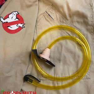May include: A yellow plastic tube with a black connector and a pink plastic mouthpiece. The tube is coiled and lying on a tan fabric background. The fabric has a red and white patch with a ghost and the words "PROPSMITH" in black.