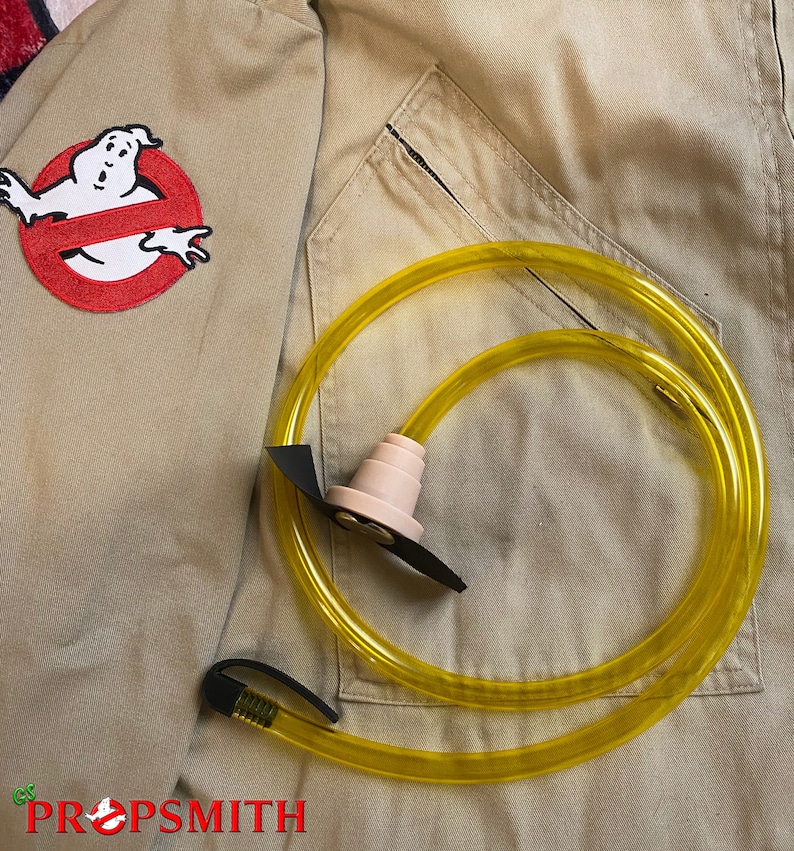 May include: A yellow plastic tube with a black connector and a pink plastic mouthpiece. The tube is coiled and lying on a tan fabric background. The fabric has a red and white patch with a ghost and the words "PROPSMITH" in black.
