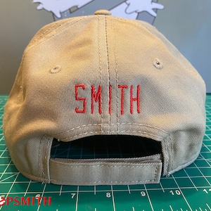 Op de afbeelding: Een beige baseballpet met een rood geborduurd "SMITH" op de achterkant.