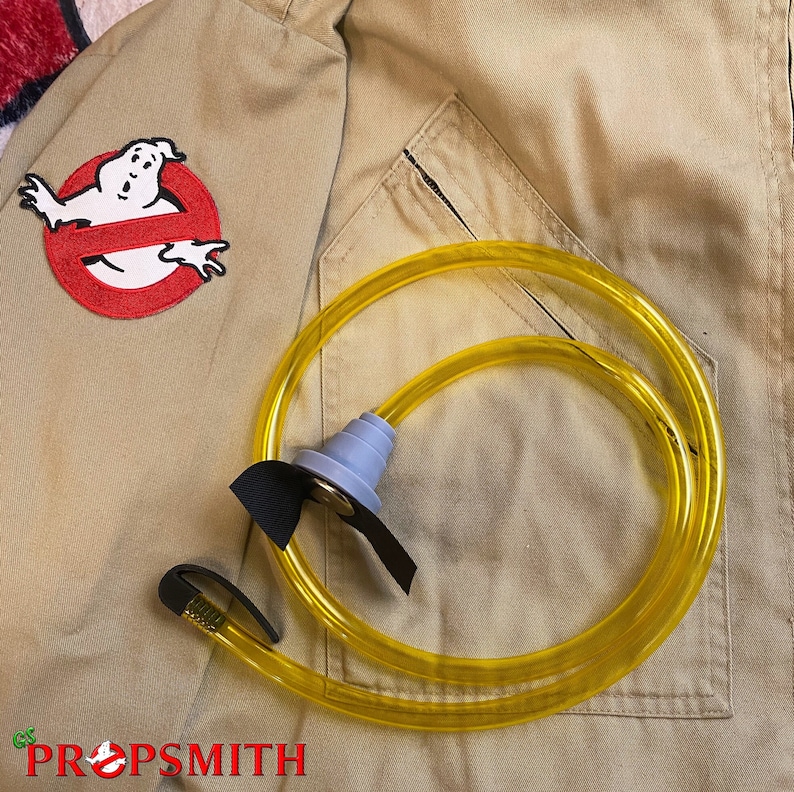 May include: A yellow plastic tube with a black handle and a gray plastic nozzle. The tube is lying on a tan fabric background with a red and white Ghostbusters logo patch.