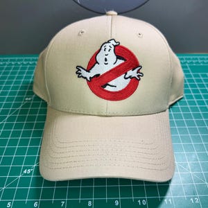 Pode incluir: Boné de beisebol bege com o logotipo dos Ghostbusters bordado. O logotipo é um fantasma branco dentro de um círculo vermelho com uma linha vermelha. O boné tem uma viseira curva e um botão.