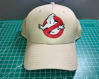 Op de afbeelding: Beige baseballpet met het Ghostbusters-logo. Het logo is een witte geest in een rode cirkel met een rode streep erdoor. De pet heeft een gebogen klep en een knoop.