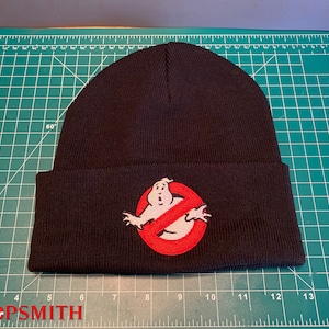May include: Black knit beanie featuring the Ghostbusters logo patch. The patch shows the Ghostbusters logo, a white ghost inside a red circle with a red slash through it. The beanie has the text "PROPSMITH" at the bottom.