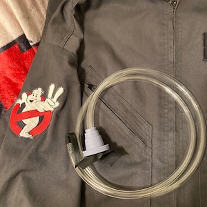 May include: A black jumpsuit with a ghostbusters logo patch on the sleeve and a clear plastic tube with a gray mouthpiece. The jumpsuit has a zipper and a pocket on the chest. The text "GS PROPSMITH" is visible on the bottom right corner of the image.