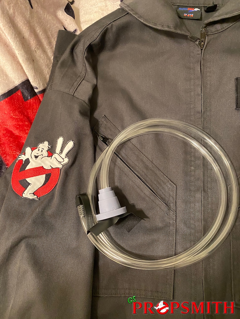 May include: A black jumpsuit with a ghostbusters logo patch on the sleeve and a clear plastic tube with a gray mouthpiece. The jumpsuit has a zipper and a pocket on the chest. The text "GS PROPSMITH" is visible on the bottom right corner of the image.