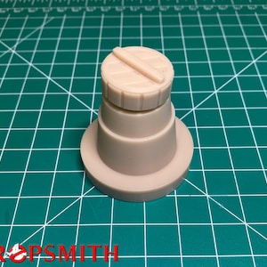 May include: A beige plastic model of a cylindrical object with a flat top and a raised, horizontal line across the top. The object is sitting on a green cutting mat.