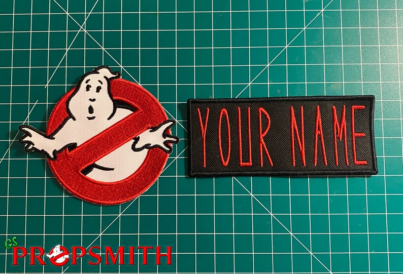 Pu&ograve; includere: Una toppa ricamata rossa e bianca con un fantasma in un simbolo di divieto, e una toppa nera con il testo "YOUR NAME" in ricamo rosso.