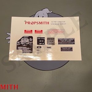May include: A sheet of stickers for Ghostbusters fans. The sheet includes a variety of stickers, including a diagram of the Ecto Goggles, a warning label, and a sticker that says "Spectral Detection Goggles Model No. E58M1 - AMPS 61 SERIAL NO. 1078475".