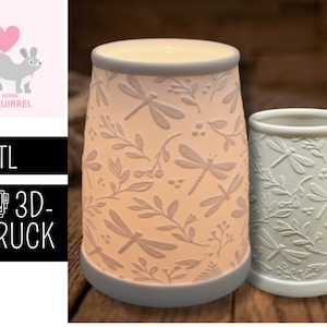 May include: Two ceramic candle holders with dragonfly and leaf patterns. The larger holder is lit from within, emitting a warm glow. The smaller holder is unlit. Both have a light blue trim and are on a wooden surface.