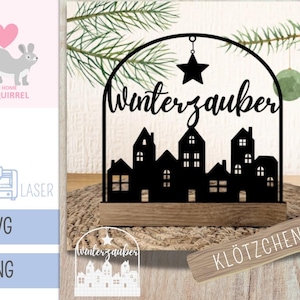 Op de afbeelding: Een zwart metalen Winterzauber-bord met een silhouet van een stad met huizen en een ster, gemonteerd op een houten voet. Het bord is ongeveer 15 cm breed en 13 cm hoog. Het ontwerp bevat de tekst "Winterzauber".