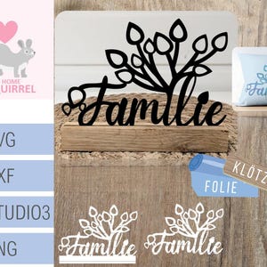 Könnte beinhalten: Schwarze Silhouette eines Baumes mit Blättern und dem Wort "Familie" darunter. Der Baum steht auf einem Holzsockel. Im Hintergrund befindet sich ein weißes Kissen mit dem Wort "Familie" darauf.