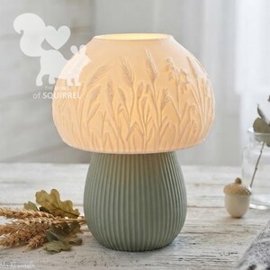 May include: A mushroom-shaped table lamp with a white translucent shade featuring embossed wheat stalks and a sage green ribbed base. The lamp emits a soft glow, and the shade displays a detailed texture of tall grasses and grain stems.