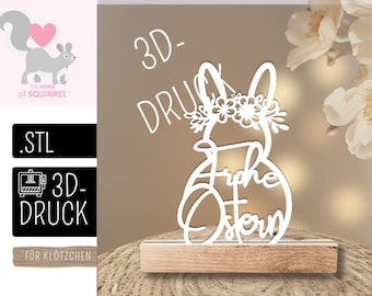 May include: A white laser-cut Easter bunny decoration with a floral crown and the words "Frohe Ostern". The bunny is mounted on a wooden base. The background is a neutral colour.