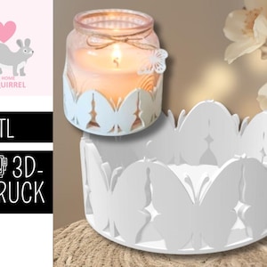 Può includere: Una candela accesa in un barattolo di vetro con un supporto bianco a forma di farfalla. Un supporto separato a forma di farfalla bianca è posto sotto. L'immagine include anche un logo di scoiattolo con il testo ".STL 3D-DRUCK".