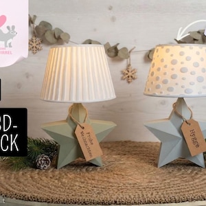 May include: Two star-shaped table lamps with light-colored bases and fabric shades. The left lamp has a pleated white shade, while the right lamp has a white shade with gray polka dots. Each lamp features a brown paper tag tied to the base.