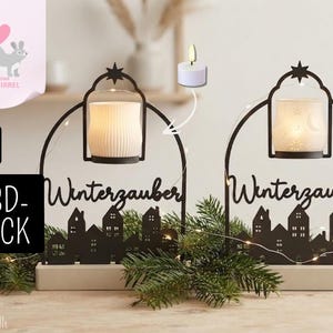 Può includere: Due portacandele in metallo nero a forma di arco con la sagoma di un villaggio e il testo tedesco Winterzauber. Ogni supporto include una tazza in ceramica bianca per lumini e una stella sulla parte superiore su una base rettangolare.