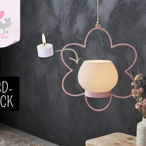 May include: A hanging light fixture featuring a pink flower-shaped metal frame and a white ribbed, rounded lampshade. A small white tea light candle is shown to the left with an arrow pointing toward the centre of the flower frame.