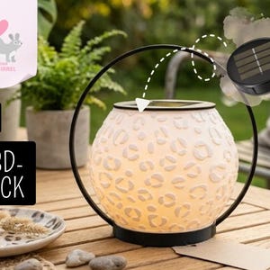 Könnte beinhalten: Eine solarbetriebene Laterne mit einer weißen, gemusterten Glaskugel und einem schwarzen Metallrahmen. Der Globus hat ein leopardenartiges Design. Die Laterne steht auf einem Holztisch, mit einem Solarpanel und einer kleinen Kerze.